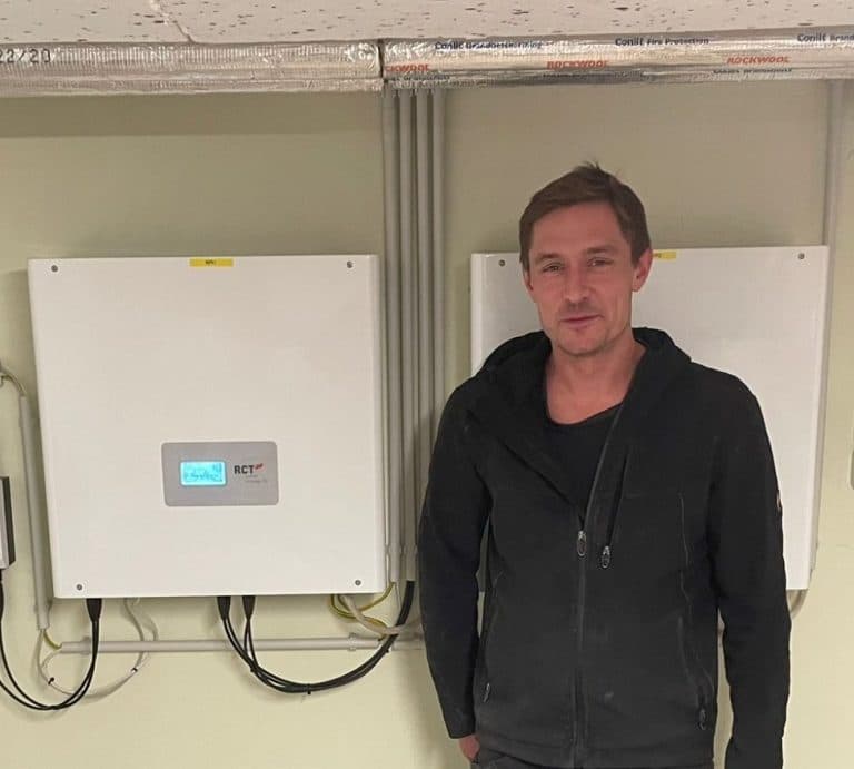 Elektromeister steht im Technikraum vor montiertem Wechselrichter und Batteriespeicher