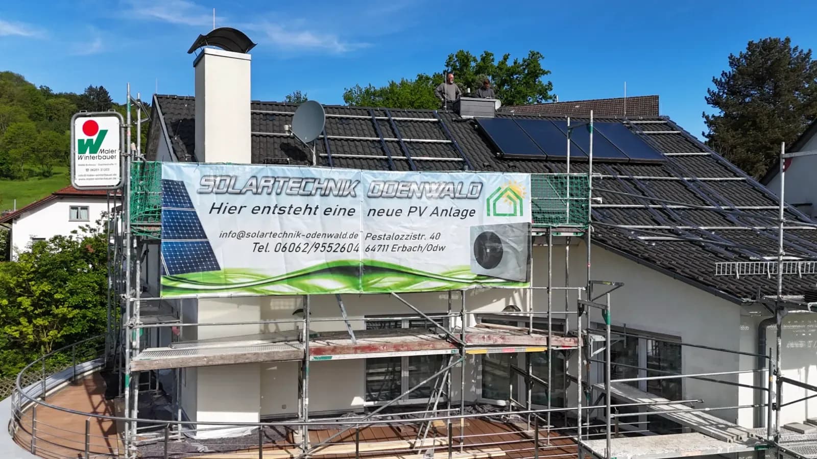 Gerüst mit Banner „Hier entsteht eine neue PV-Anlage" vor einem Haus, zwei Monteure arbeiten am First