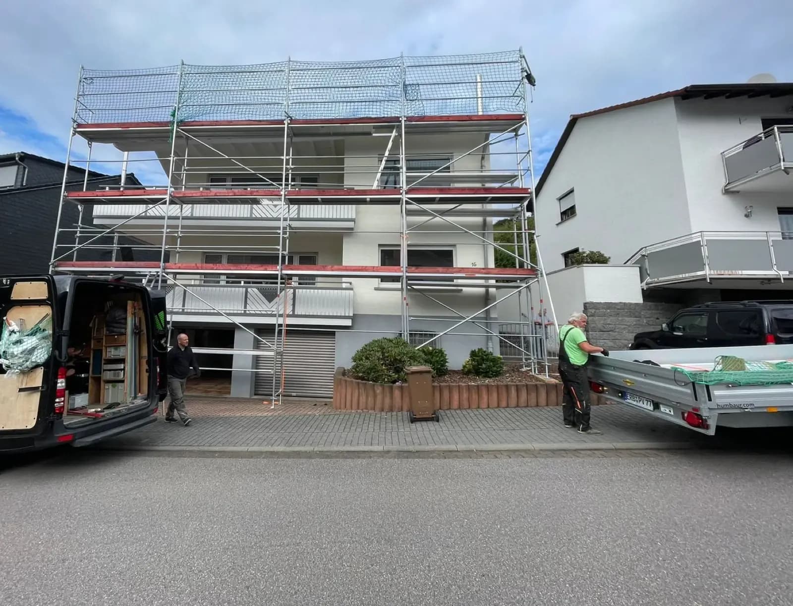 Aufbau eines dreistöckigen Gerüsts vor einem Mehrfamilienhaus, daneben ein beladener Anhänger