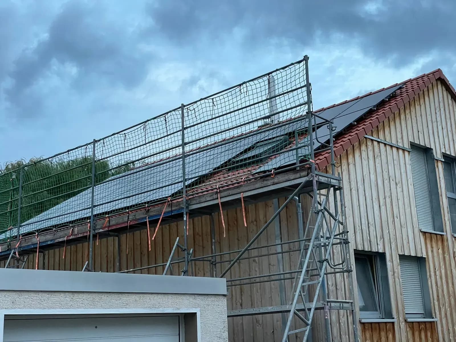 Holzverkleidetes Haus in der Dämmerung, Gerüst mit Netz vor dem fast fertigen Modulfeld